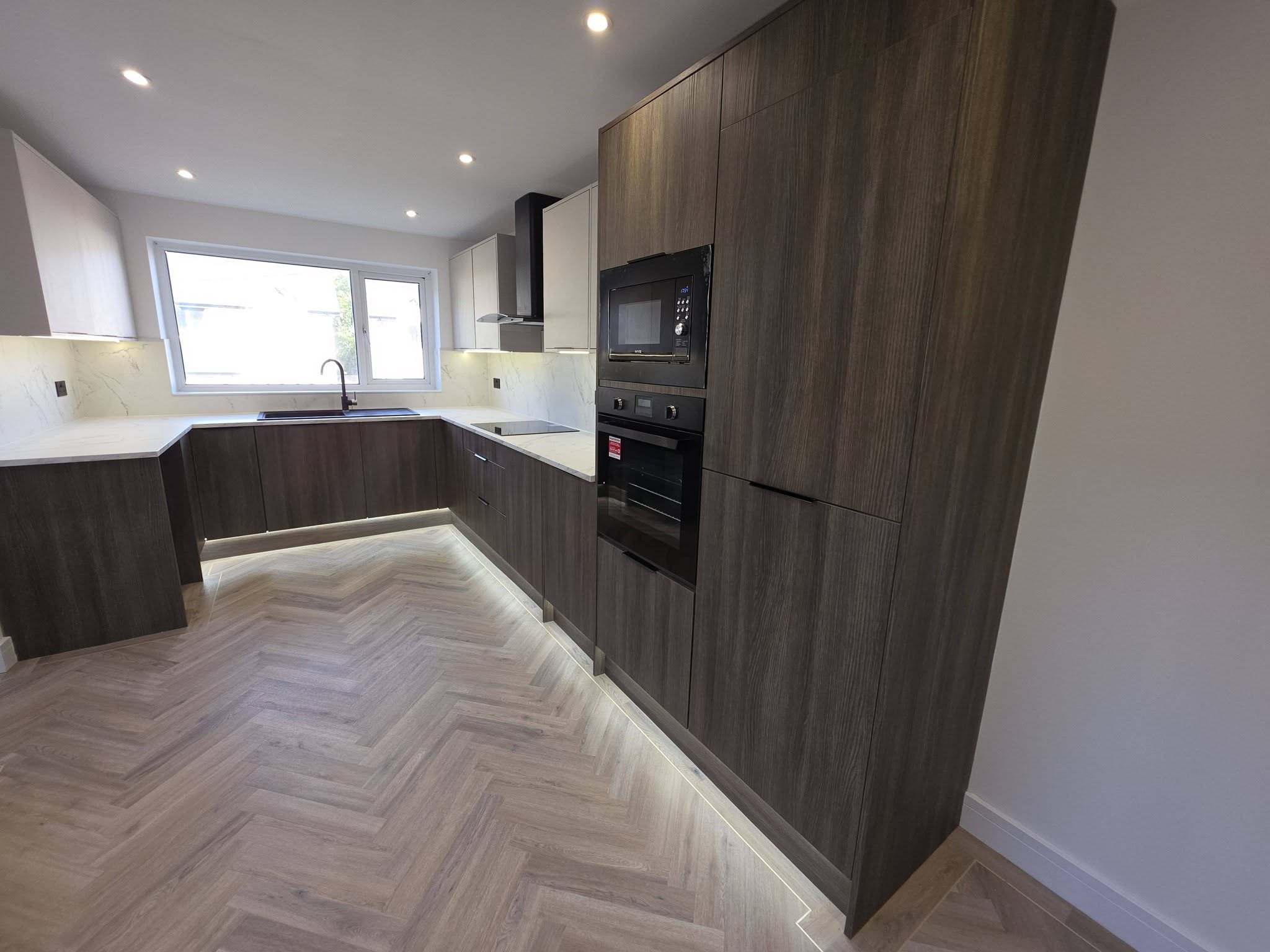Brand-new modern kitchen with dark wood-effect and cream units, integrated ovens, plinth lighting and herringbone LVT flooring